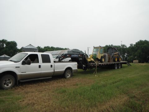 2000 Ford F-250 - A white Ford F-250 truck tows a trailer loaded with a skid steer loader across a grassy field.