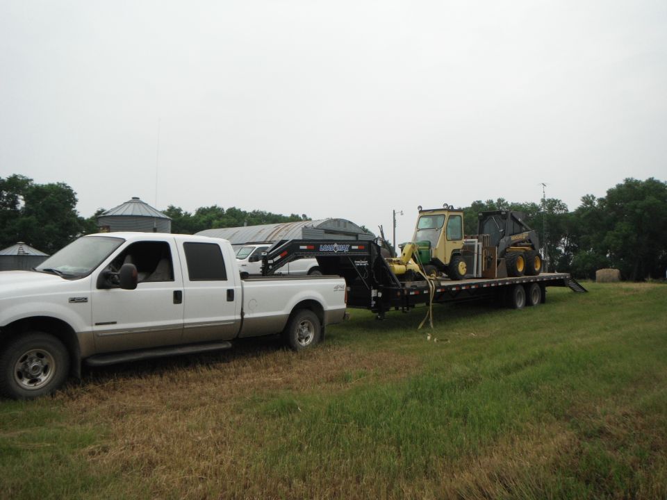 White Ford F-250 truck, tan lower body, towing a flatbed trailer with a yellow skid steer loader. Cloudy sky