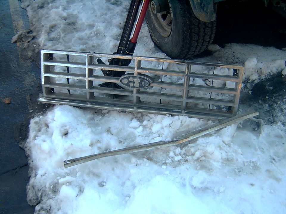 Ford grille with oval emblem resting on snow beside a chrome trim strip, tire behind.