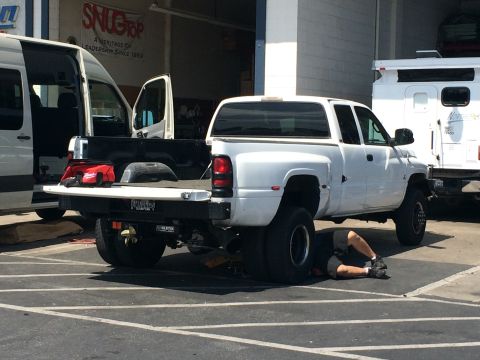 1997 Dodge 3500 - A mechanic works on the underside of a white 1997 Dodge 3500 dually pickup truck parked in a lot.