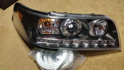 2003 Ford Crown Victoria - Aftermarket black-housing projector headlight with dual halo rings, an amber turn-signal lens, and a row of LED accents, with wiring on the…
