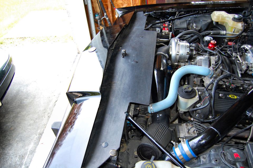 Engine bay with supercharger, blue hose, and air intake. Part of a car's fender is visible on the left