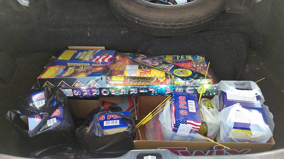 Trunk packed with boxes and bags of fireworks, including sparklers and larger display fireworks. A spare tire is visible above