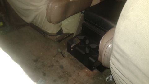 1996 Ford Bronco - The interior of a vehicle shows a black center console with a dual cup holder assembly. Tan seats and the floor are visible.