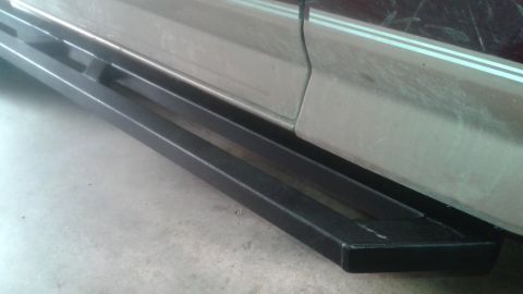 1996 Ford Bronco - A close-up, side-angle view of a black metal nerf bar installed below the rocker panel of a light-colored SUV. The nerf bar features a rect…