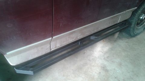 1996 Ford Bronco - The side of a maroon SUV with a custom black metal step rail installed below the rocker panel. The vehicle appears to be parked on a concre…