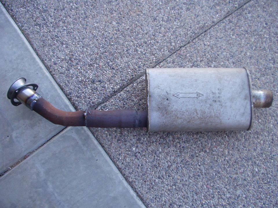 Removed oval muffler with curved inlet pipe and flange lying on concrete sidewalk