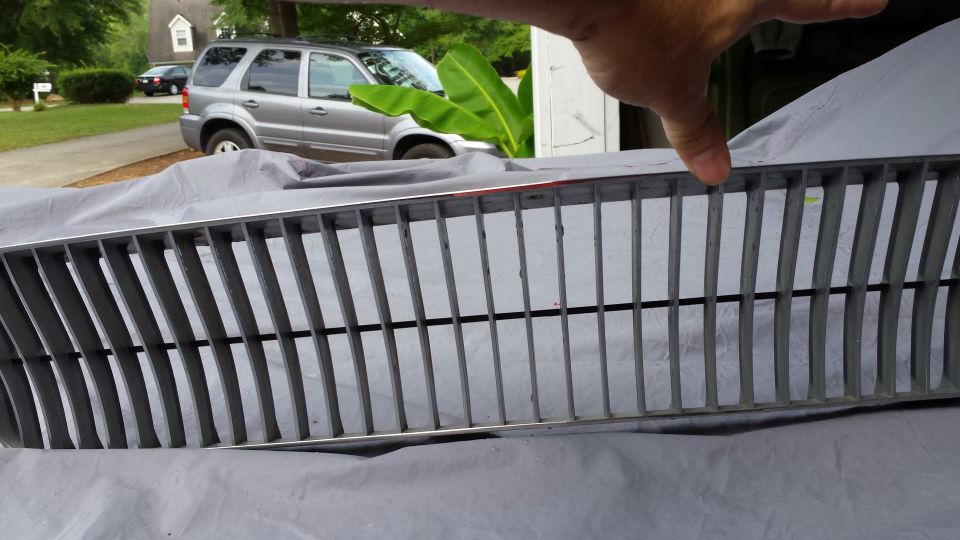 Gray car grille with vertical slats, silver SUV in background.
