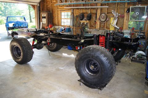 1979 Ford Bronco - The chassis and suspension of a classic Ford Bronco are shown in a garage setting. The vehicle is partially disassembled with large off-roa…