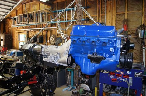 1979 Ford Bronco - A blue V8 engine and transmission assembly are suspended by chains in a garage workshop, above a partially assembled vehicle frame.