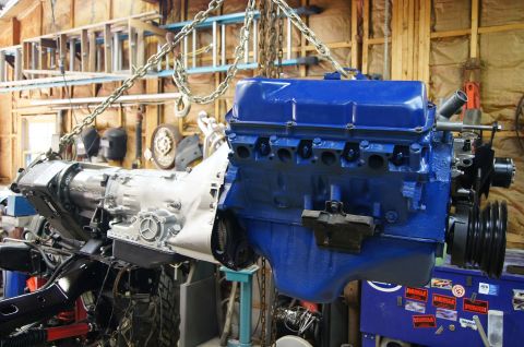 1979 Ford Bronco - A blue Ford V8 engine and silver transmission are suspended by chains in a garage setting. The engine is mounted on a black frame with visi…