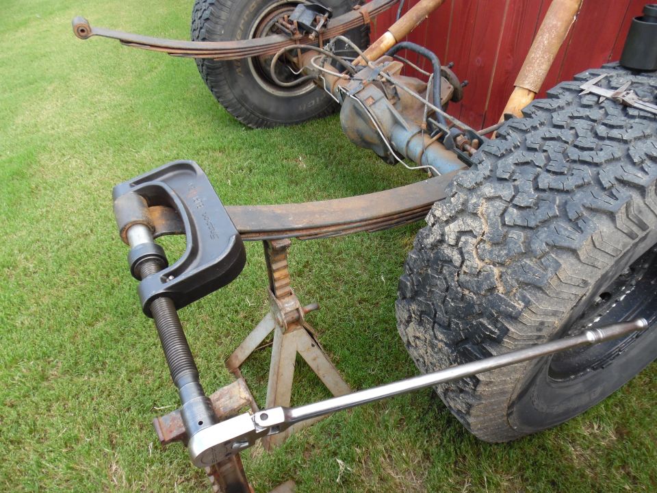 C-clamp press and breaker bar working on a rusty leaf spring near an off-road tire
