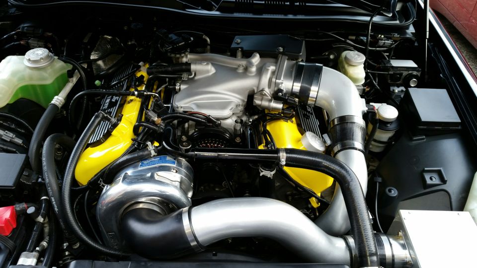 Engine bay with supercharger, yellow valve covers, and custom intake piping.
