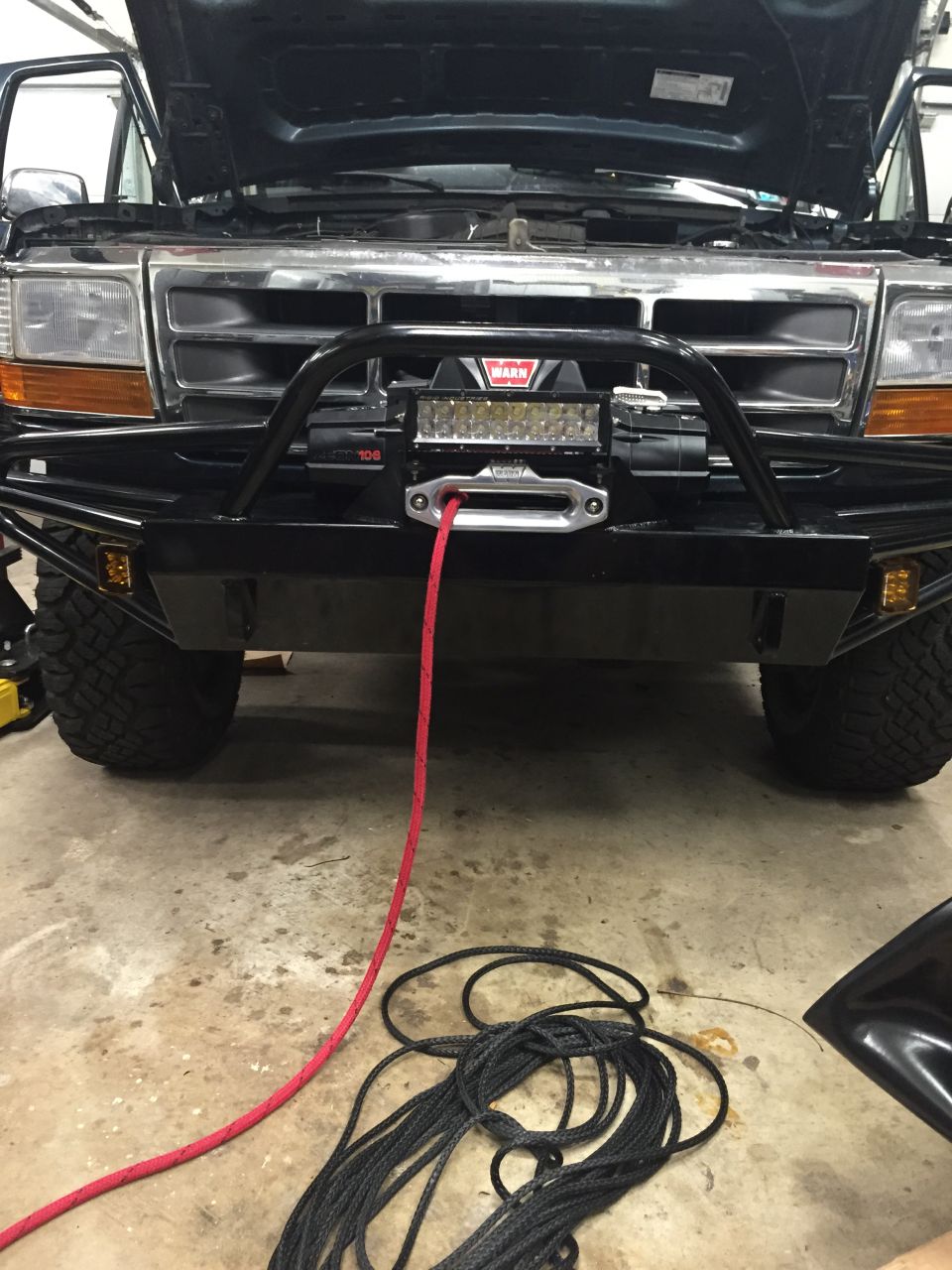 Front of a Ford F-Series truck with hood open, custom bumper, and winch. Red winch line on the ground