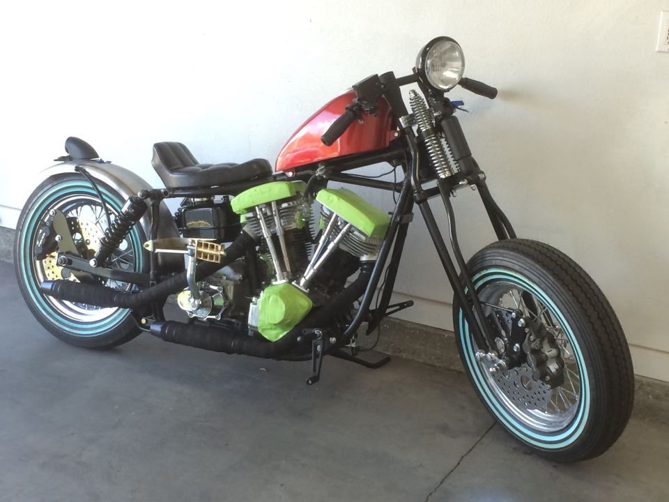 Chopper motorcycle with red fuel tank, black seat, and green engine covers. Whitewall tires and chrome exhaust are visible