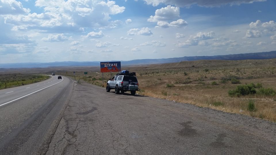 White SUV parked on roadside with Utah welcome sign in background. Roof rack loaded with gear