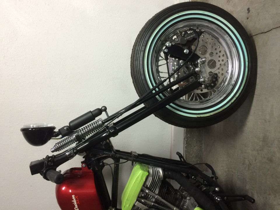 Motorcycle front end with black handlebars, dual springs, and a chrome wheel with a whitewall tire. Red fuel tank visible