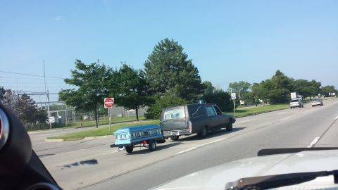 1995 Ford Bronco - A hearse tows a coffin-shaped trailer down the road on a sunny day.