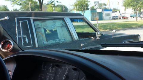 1995 Ford Bronco - A dark-colored hearse is seen from the interior of another vehicle, with a website sticker on its side.