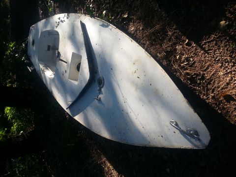 1973 Ford F-100 - A white boat hull rests on the ground, surrounded by foliage and debris. A dark fin is visible on its surface.