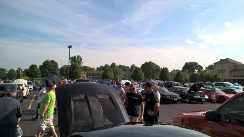 2004 Ford Crown Victoria - People mill around a parking lot filled with cars, some with their hoods open, under a partly cloudy sky.