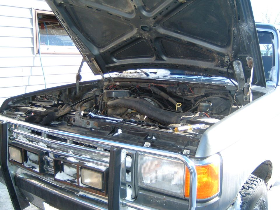Silver Ford truck with raised black hood showing engine bay, brush guard, in snowy yard