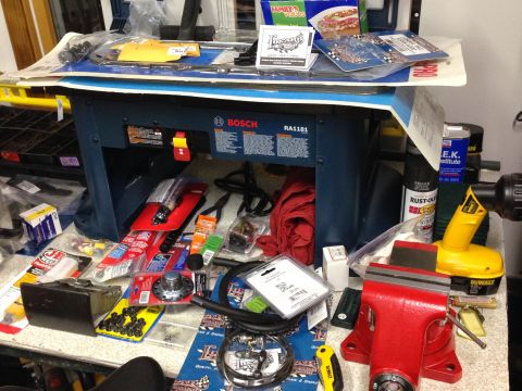 1979 Ford Bronco - A workbench is covered in various tools and parts, including a blue Bosch RA1181 table saw, a red vise, a yellow DeWalt drill, and several…