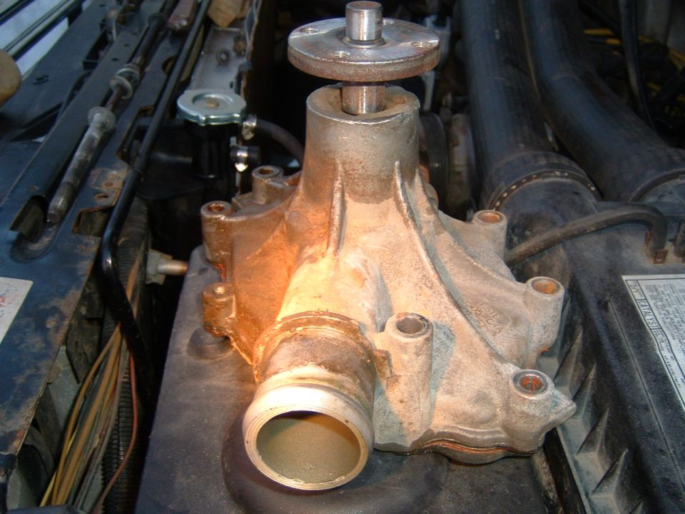 Close-up of a water pump assembly with a shaft protruding from the top, situated within an engine bay