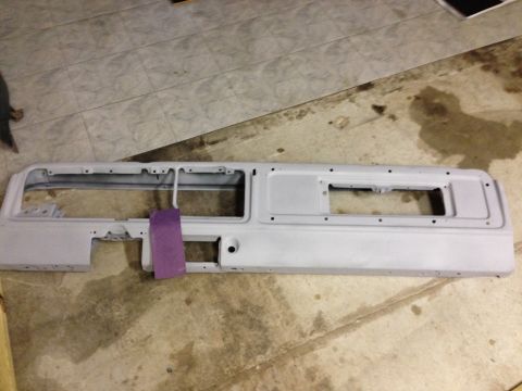 1979 Ford Bronco - A gray, primed dashboard panel with two large rectangular cutouts is laid flat on a concrete floor. A purple sanding block is attached to t…