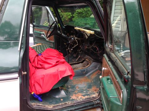 1979 Ford Bronco - The interior of a vintage green Ford Bronco with the driver's door open, showing a red tarp covering the passenger seat and the dashboard r…