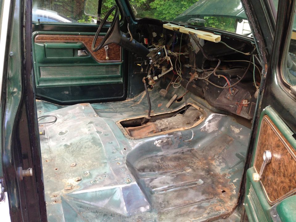 Interior of a vintage Ford Bronco with exposed, rusty floor pan, dashboard, steering wheel, and wiring.