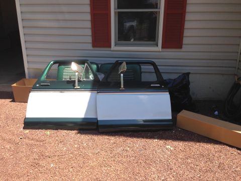 1979 Ford Bronco - Two detached doors, likely from a 1979 Ford Bronco, are leaning against a house. The doors are primarily white with a dark green top and bo…