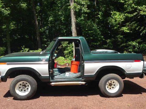 1979 Ford Bronco - The side profile of a two-tone green and silver 1979 Ford Bronco is shown with the driver's side door open, revealing the interior. An oran…