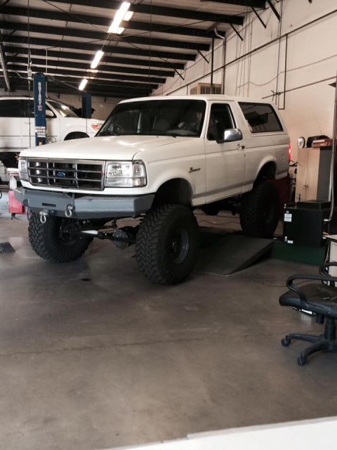 1995 Ford Bronco - A lifted, white 1995 Ford Bronco is parked in a garage with its front end elevated on a ramp. The vehicle features large off-road tires and…