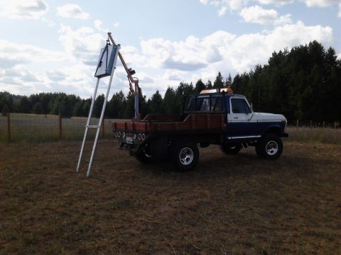1989 Ford Bronco - A blue and white Ford flatbed truck with a crane attachment is parked on a dry grassy field with trees in the background.