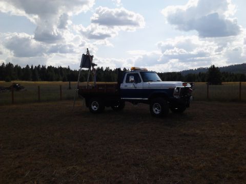 1989 Ford Bronco - A two-tone blue and white pickup truck with a flatbed is parked in a grassy field with a forest in the background. A ladder-like structure…