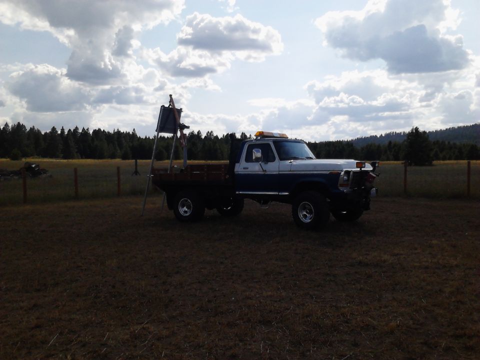 Blue and white pickup truck with a flatbed and a ladder structure parked in a field.