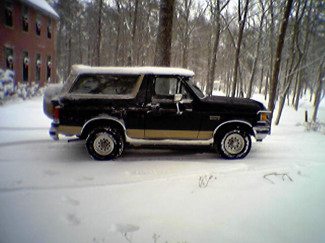 Black and tan 1990s Ford Bronco in profile parked in deep snow by a red house
