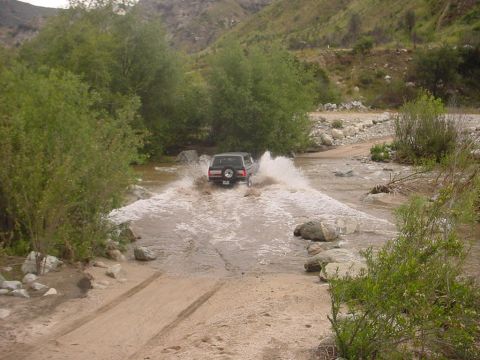 1988 Ford Bronco - An SUV navigates a river crossing on a dirt path, surrounded by lush greenery and rocky terrain.