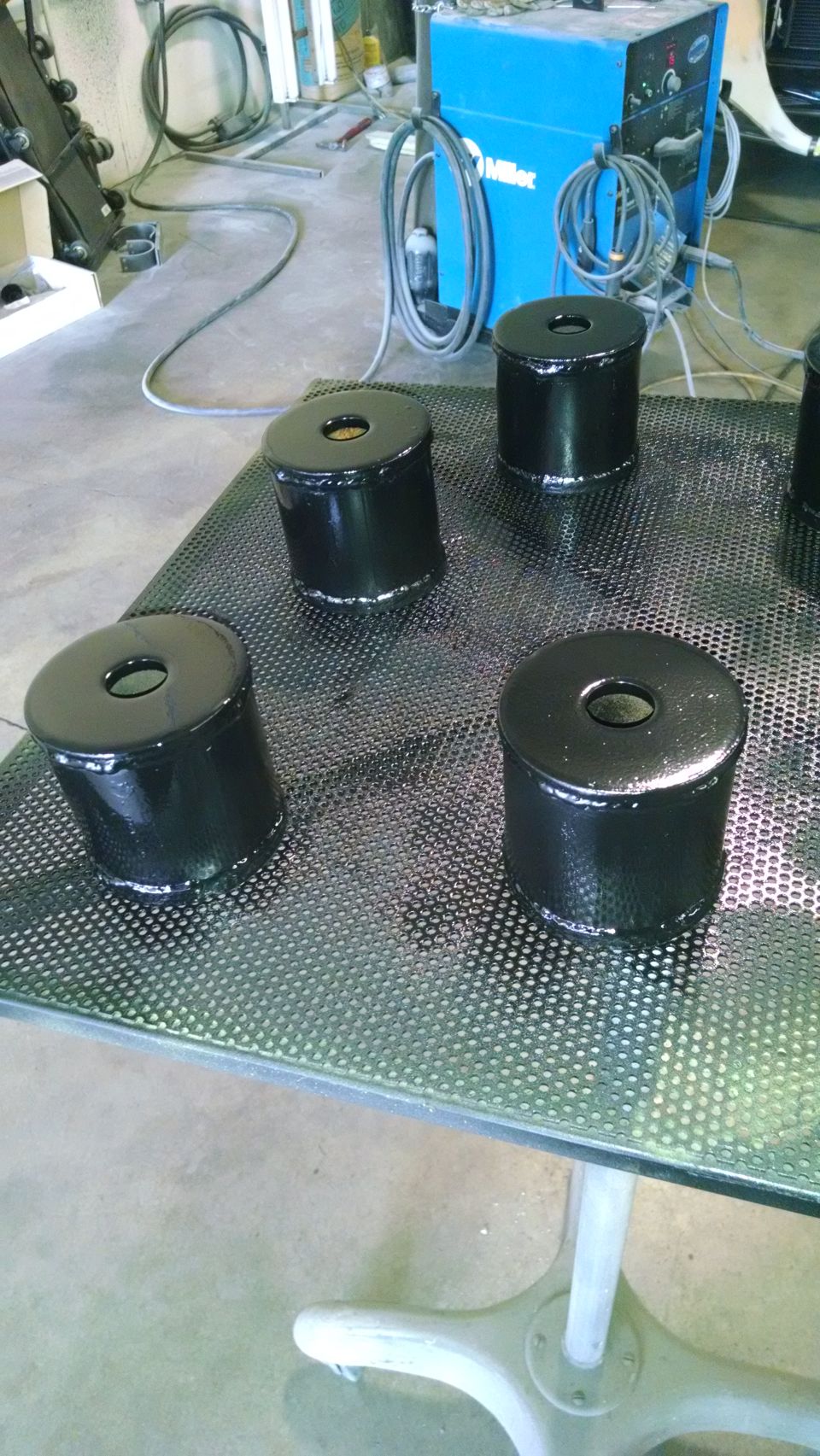 Black cylindrical metal parts on a perforated table with a welding machine in the background.