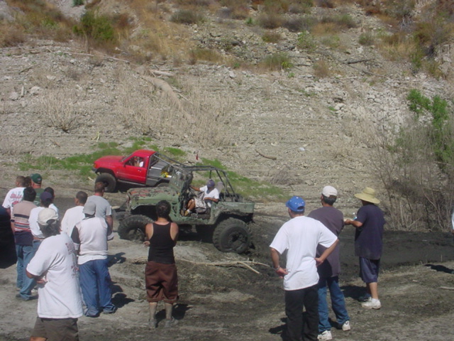 Red tube-bodied off-road truck on muddy hill with spectators watching