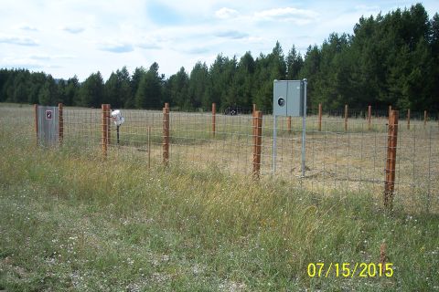 1989 Ford Bronco - A wire fence with wooden posts stands in a grassy field, with a utility box and a gate visible. A line of pine trees forms the background u…