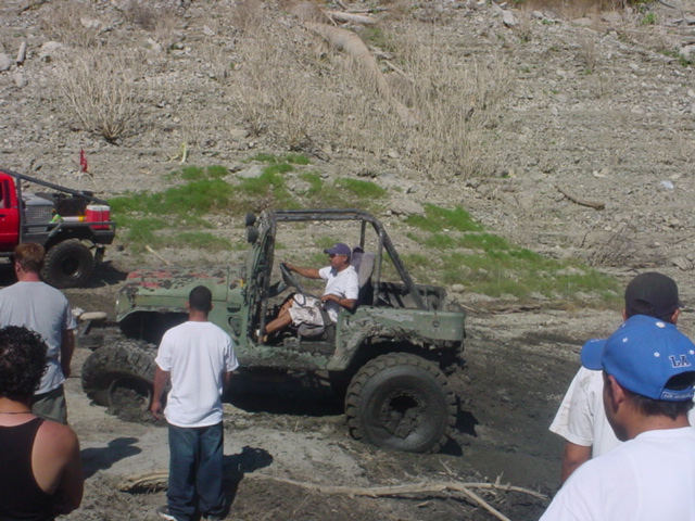 Mud-covered green Jeep Wrangler with roll cage, driver in white shirt, and large tires, stuck in mud with people watching
