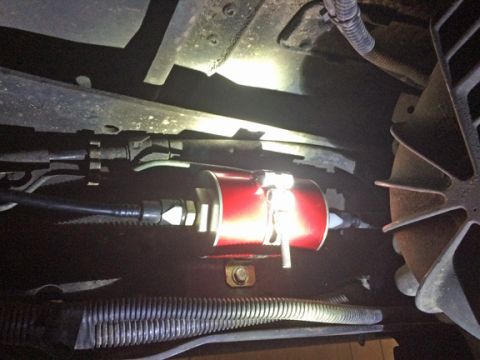 1990 Ford Bronco - A close-up view shows a red fuel filter with black hoses attached, mounted underneath a vehicle. A metal bracket and clamp secure the filte…