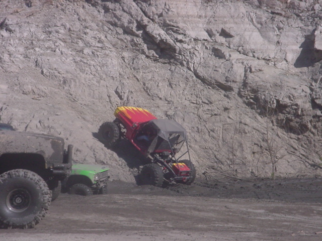 Red buggy with yellow flames climbing a muddy incline, with other off-road vehicles nearby
