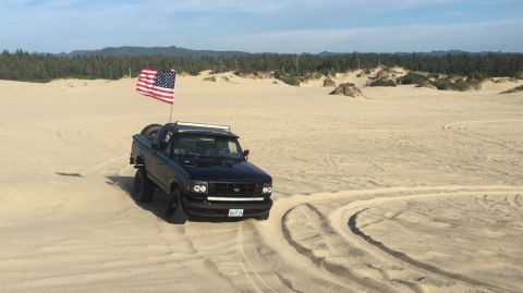 1992 Ford Bronco - A black Ford Bronco with an American flag is driving on sand dunes. The vehicle has a light bar on the roof and aftermarket headlights.