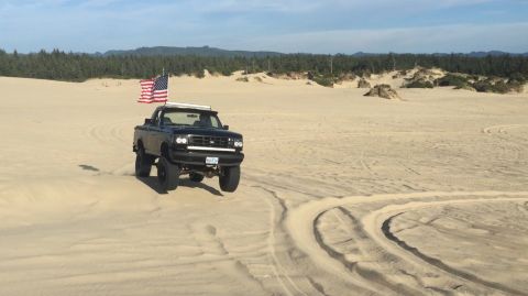 1992 Ford Bronco - A black Ford Bronco with an American flag mounted on top drives across sand dunes with a forest in the background.