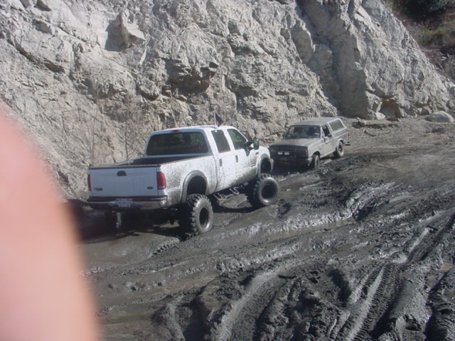 White lifted Ford pickup and white Ford Bronco driving through muddy trail below rocky cliff