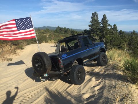 1992 Ford Bronco - A black 1992 Ford Bronco with blue pinstripes is parked on a sand dune with an American flag flying behind it. The Bronco has a spare tire…