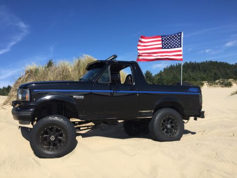 1992 Ford Bronco - A lifted black 1990s Ford Bronco with an American flag mounted on the back is parked on a sandy dune with sparse vegetation and a treeline…